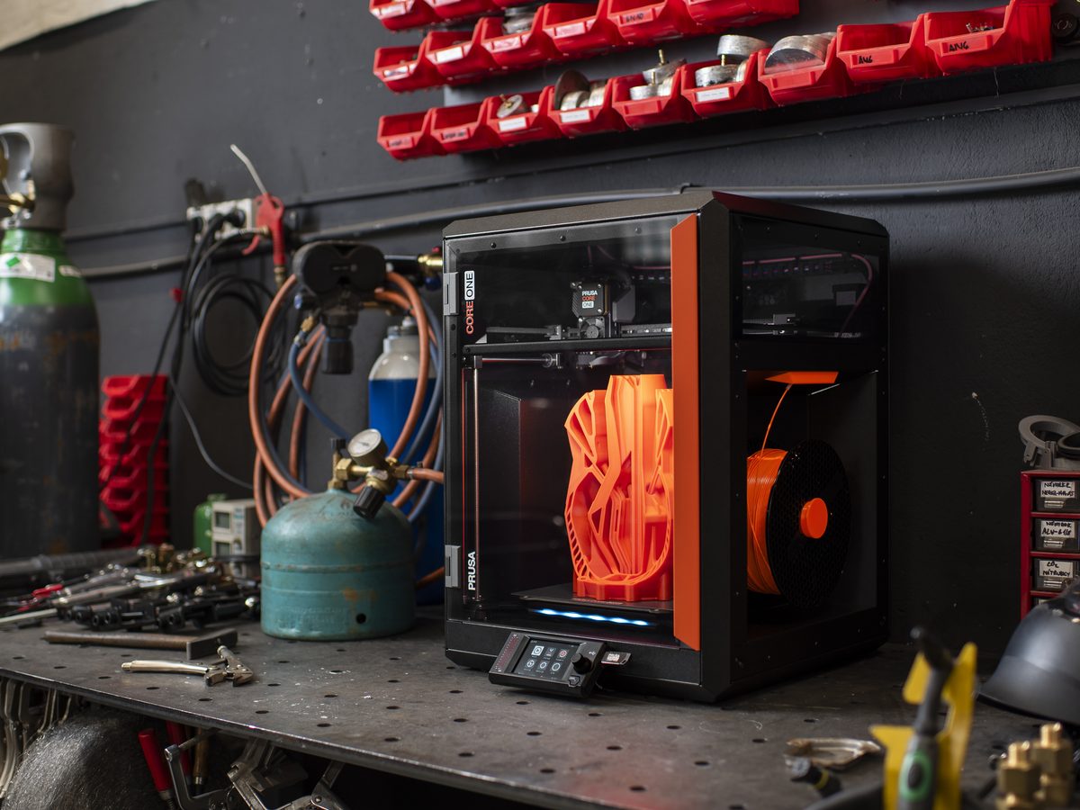 Impresora 3D Prusa CORE One sobre un banco de taller, imprimiendo una pieza naranja