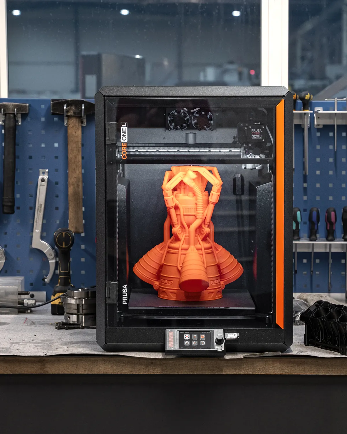 Impresora Prusa Core One L con una pieza naranja impresa dentro, sobre un banco de trabajo con herramientas