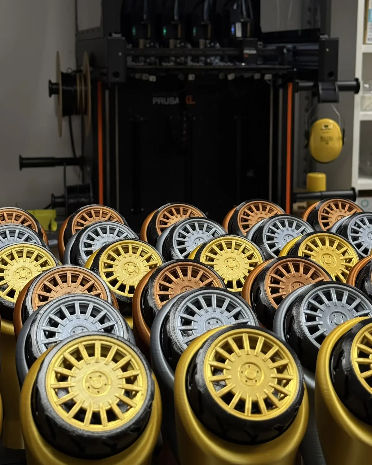 Serie de llantas impresas en 3D en dorado, plata y cobre, alineadas delante de una impresora Prusa en el taller de La Botiga 3D